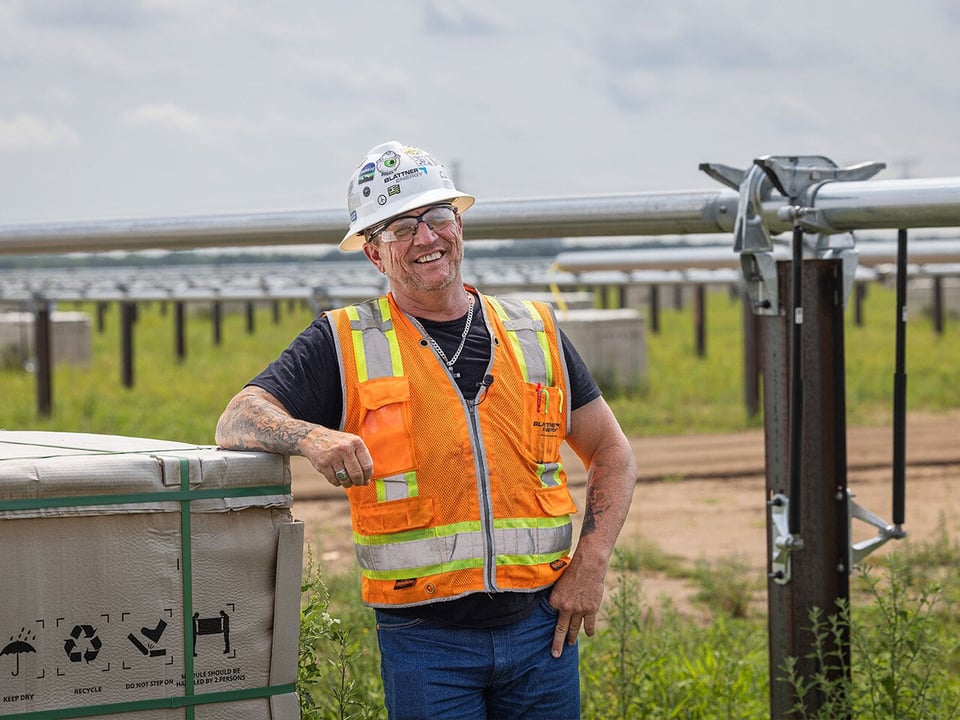 smiling male, tattooed construction worker wearing jeans, a safety vest, glasses and hardhat stands among the solar array on a solar energy project