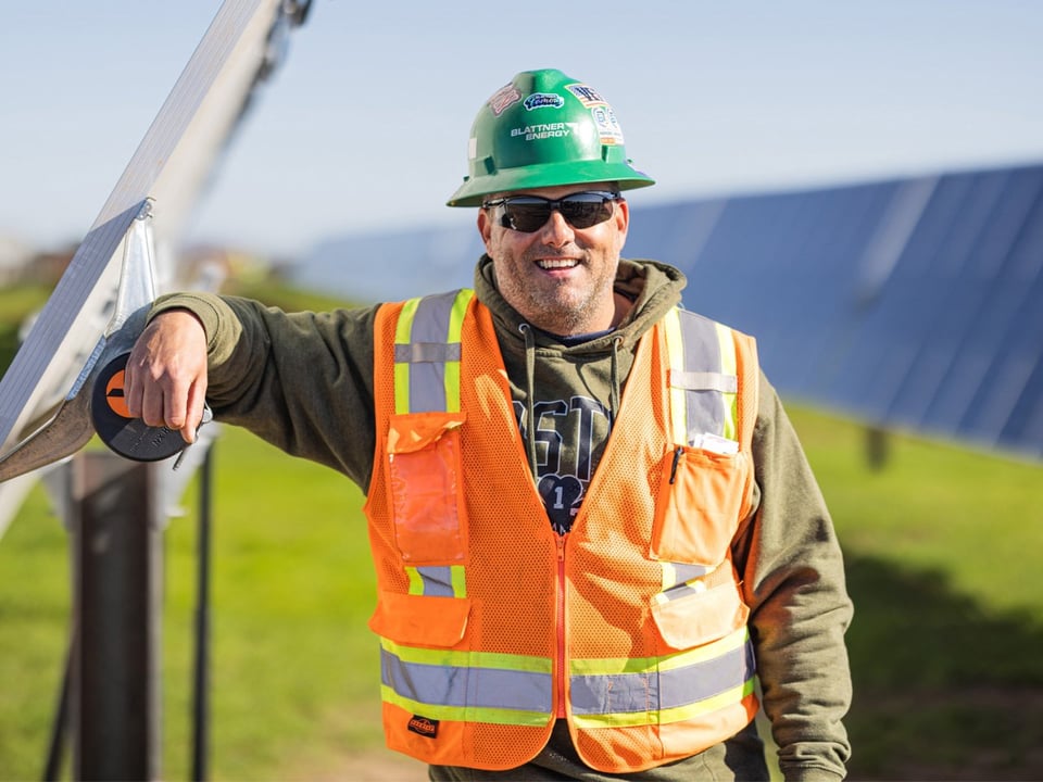 a smiling mature male construction worker in a green hardhat laughs as he poses in a solar array on a solar energy project