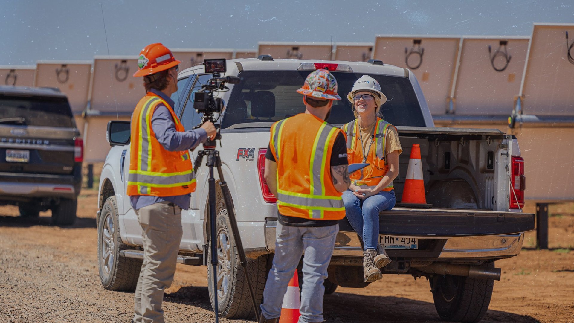 Header-2048x1152- young female construction worker being interviewed by a film crew while she_s sitting on the tailgate of a white pickup truck positioned within a utility scale solar energy project array