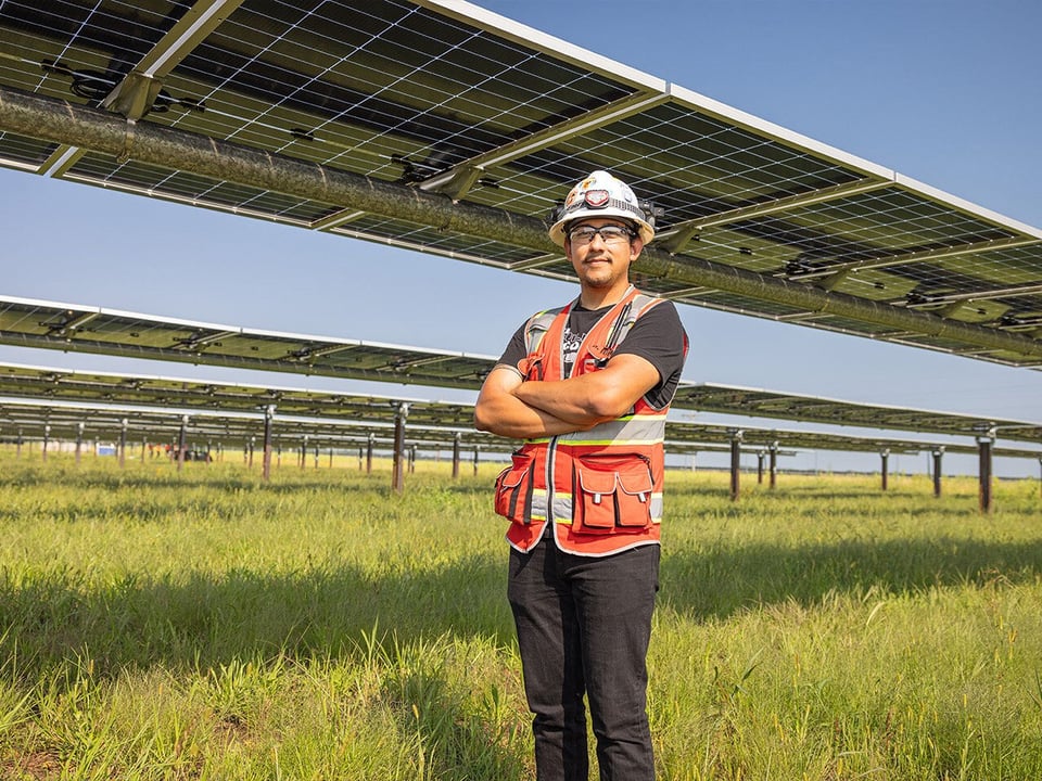 a construction worker wearing an orange safety vest and white hardhat stands proudly, arms crossed, in the shade of a solar array on a utility scale solar project in Minnesota