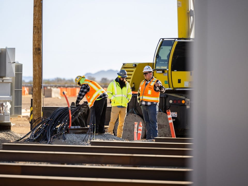 Three workers on an energy project are planning the installation of black cable on a renewable energy project with a large yellow crane in the background