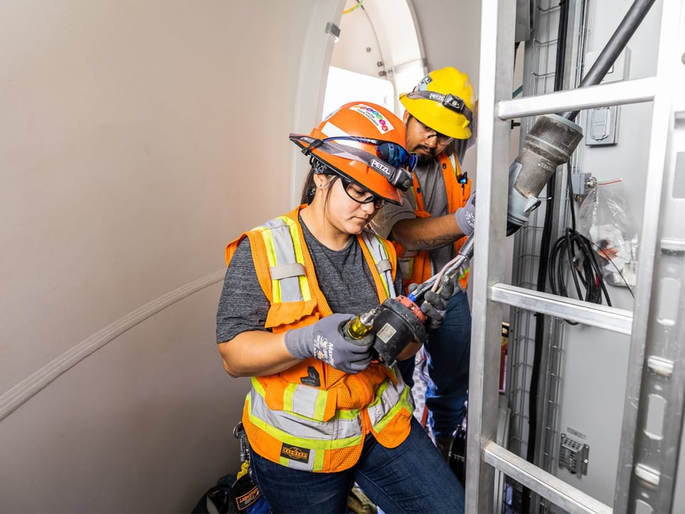 Close up of female electrician installing electrical components at the base of the interior of a wind turbine as a male coworker observes