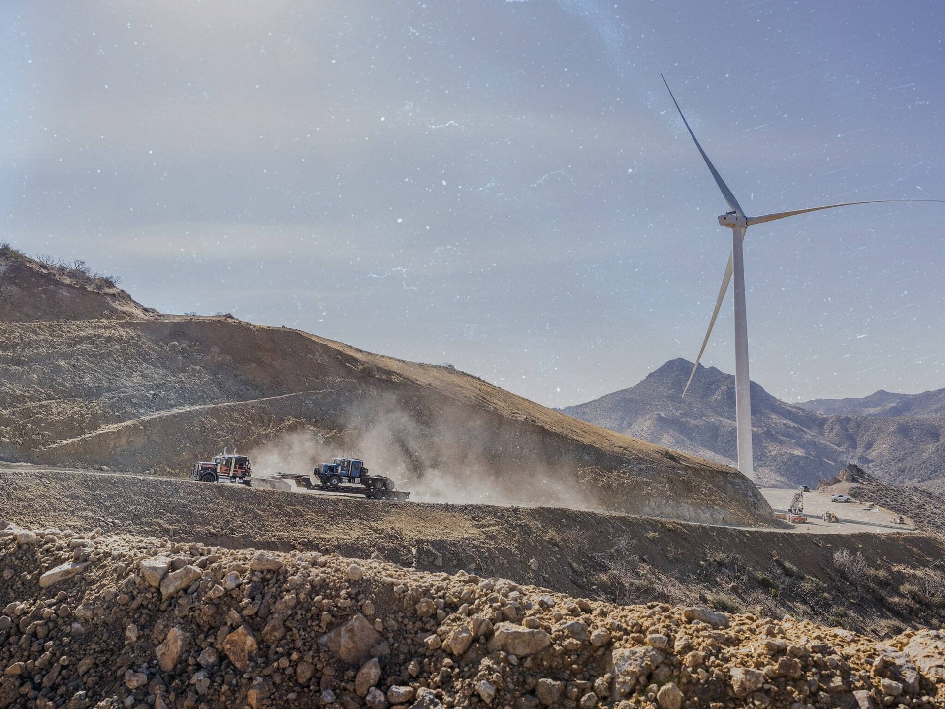 Semi truck hauling a truck up a mountain road on a rocky mountainous wind energy project with wind turbine in the background-min