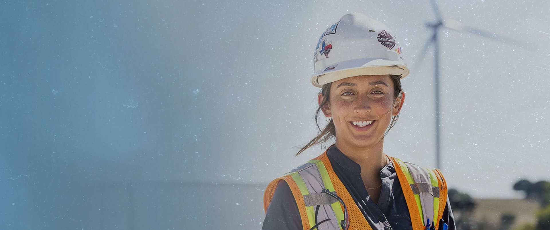 female construction worker on a wind energy project site in a white hard hat and orange safety vest looking at the camera with a tall wind turbine in the background