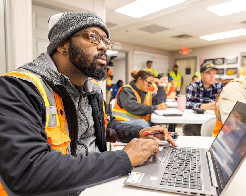 Man with laptop in forefront as others on project site gather in room.